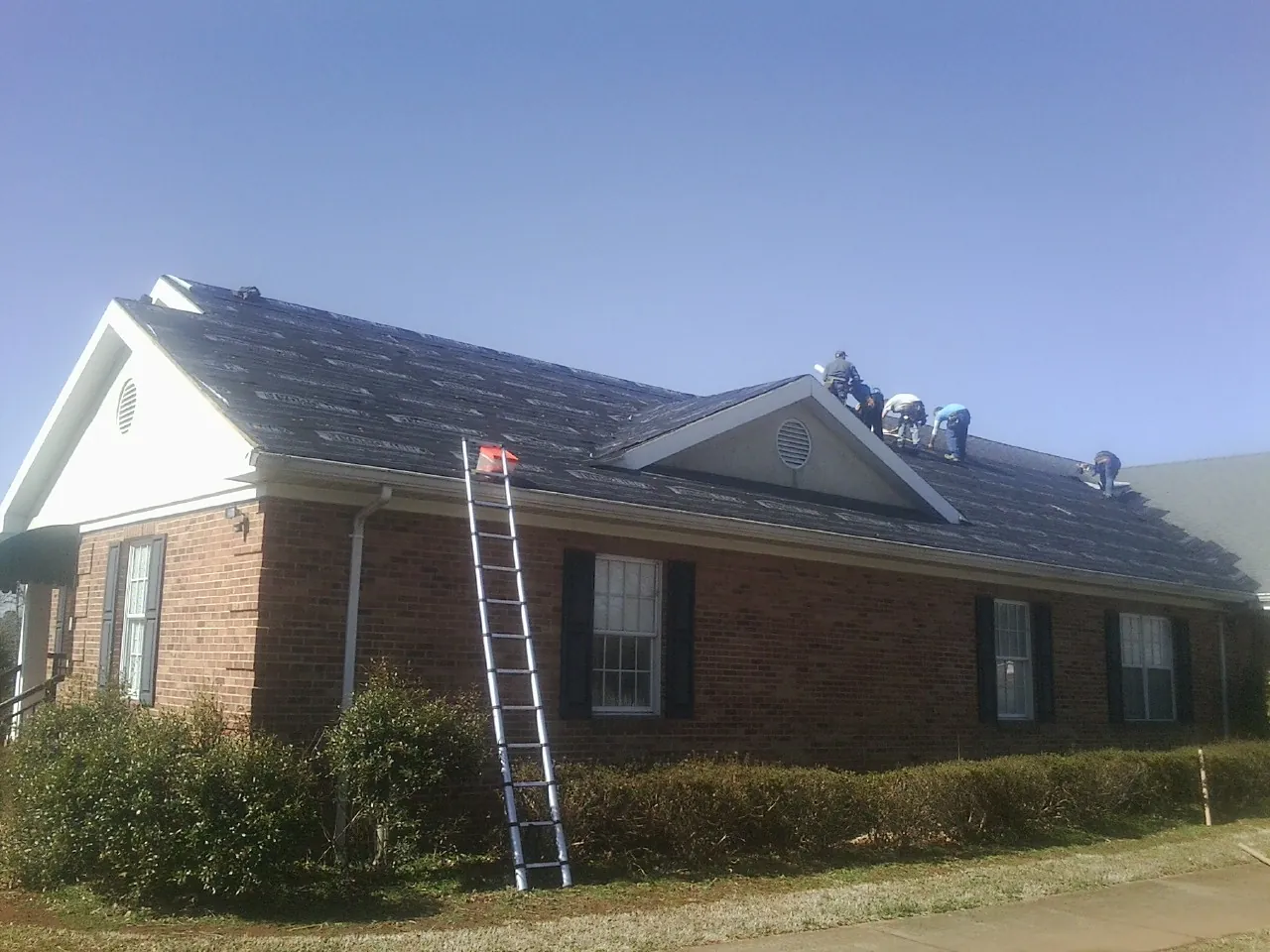 Crew tearing shingles off a brick ranch roof with a ladder set — Metal Roof Repair work by our crew in Lake George